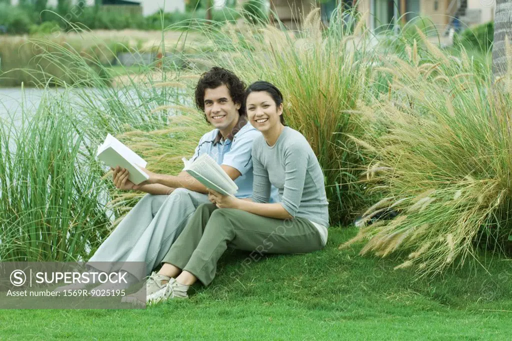 Couple sitting on grass with books, smiling at camera