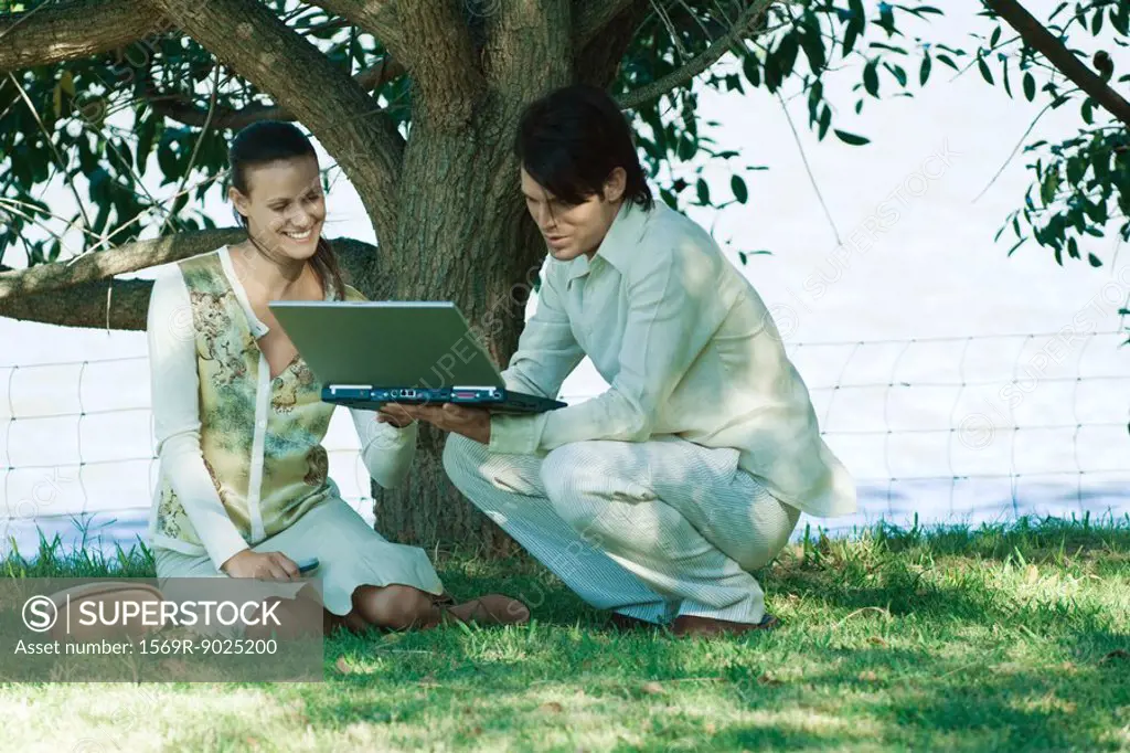 Couple under tree, man holding open laptop