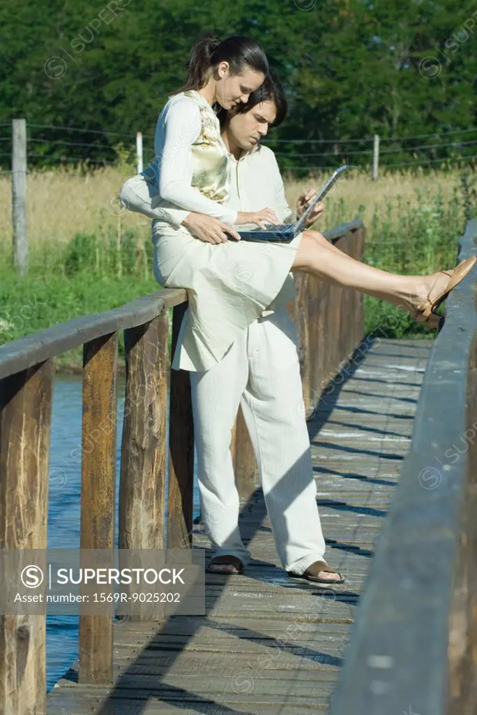 Couple on footbridge over water, woman sitting on rail, using laptop