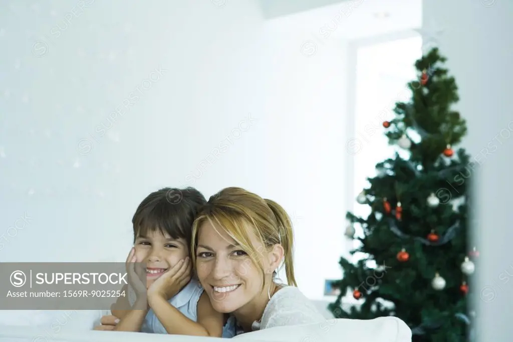 Girl and mother on sofa smiling at camera, Christmas tree in background