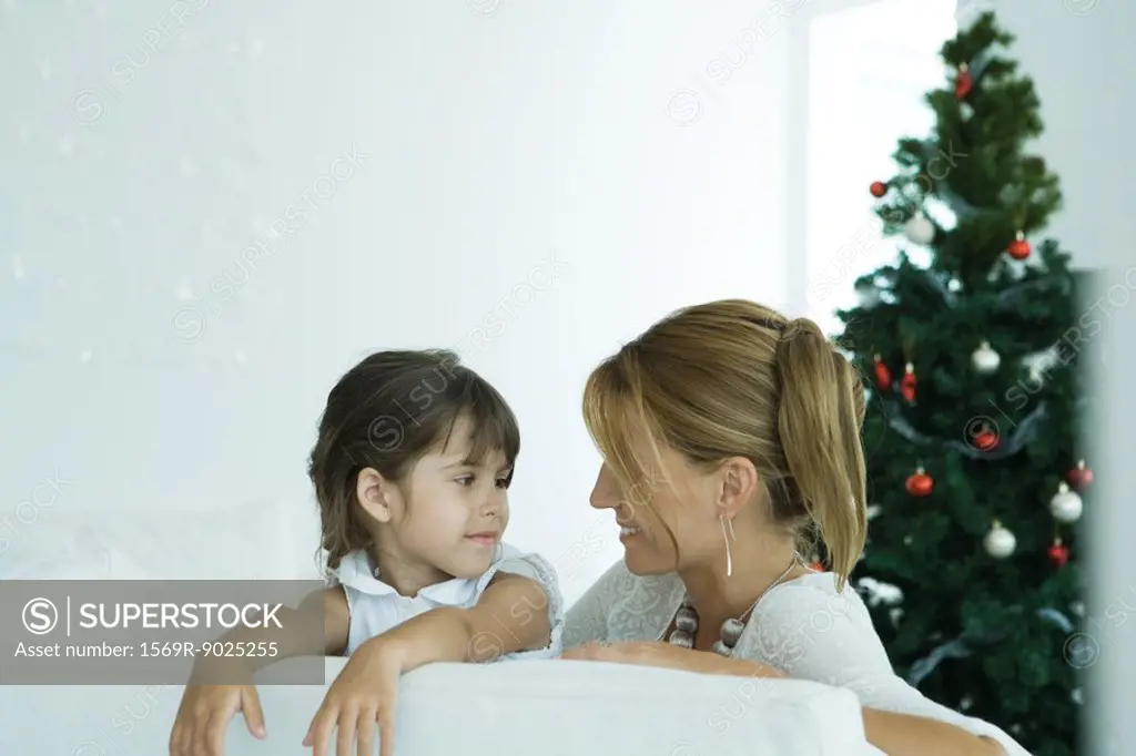 Girl and mother on sofa smiling at each other, Christmas tree in background