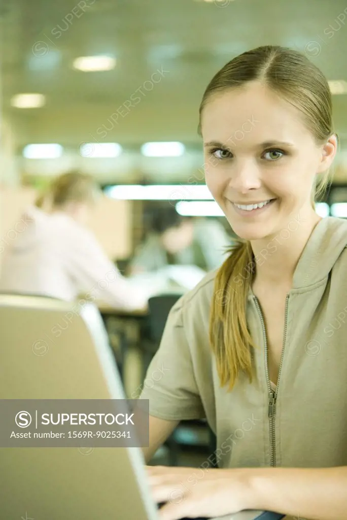 Young woman using laptop in library, smiling at camera