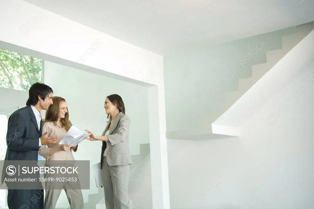 Female real estate agent showing house to young couple, pointing to blueprints