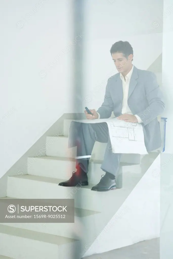Man in suit sitting on stairs, looking at blueprints, using cell phone, seen through glass