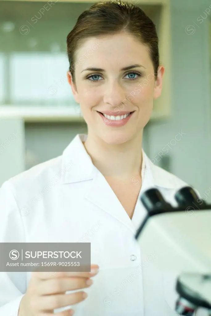 Female lab worker with microscope, holding up slide, smiling at camera, portrait
