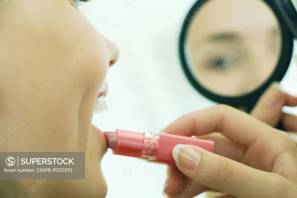 Teenage girl putting on lipstick, looking at self in hand mirror, cropped view