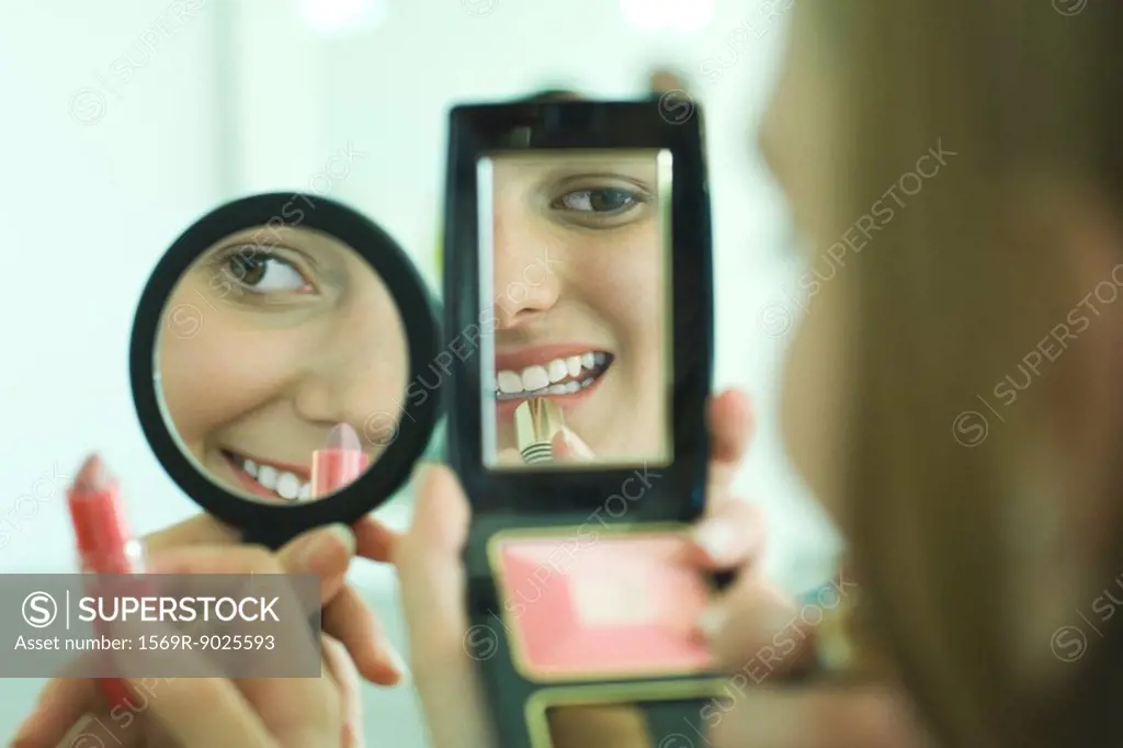 Two young friends putting on lipstick, looking at selves in hand mirrors, cropped view
