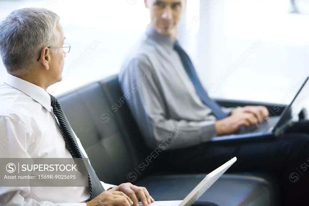 Two businessmen sitting on sofa, looking at each other, both using laptops computers