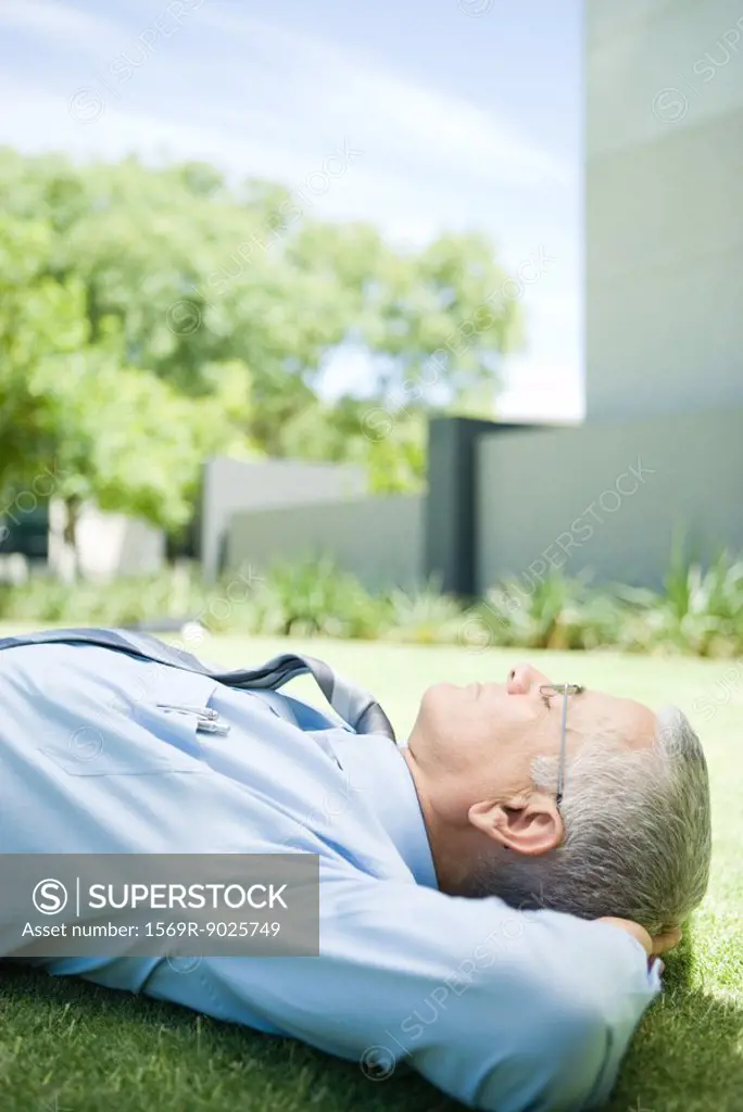 Mature businessman lying on ground outdoors, hands behind head, eyes closed, side view