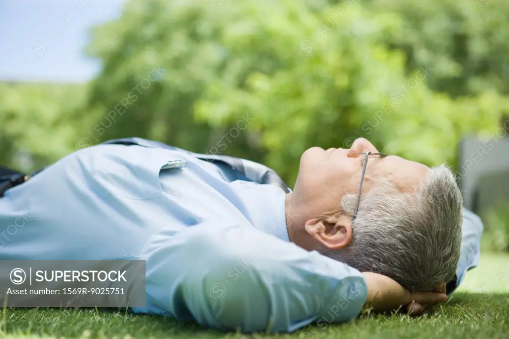 Mature businessman lying on ground outdoors, hands behind head, eyes closed, close-up