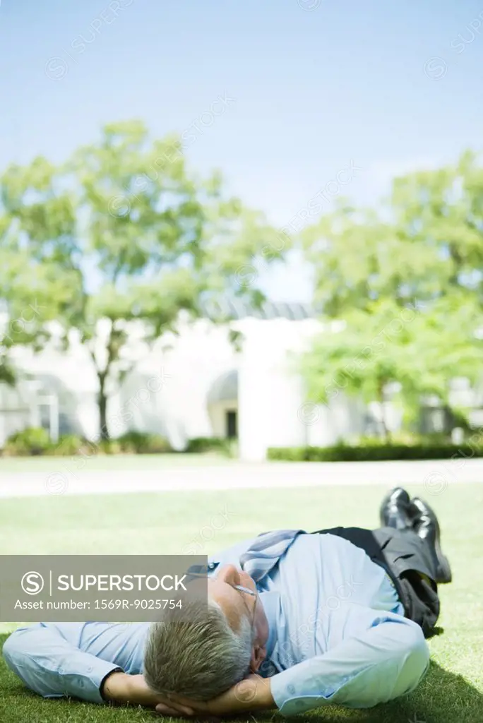 Mature businessman lying on ground outdoors, hands behind head, full length