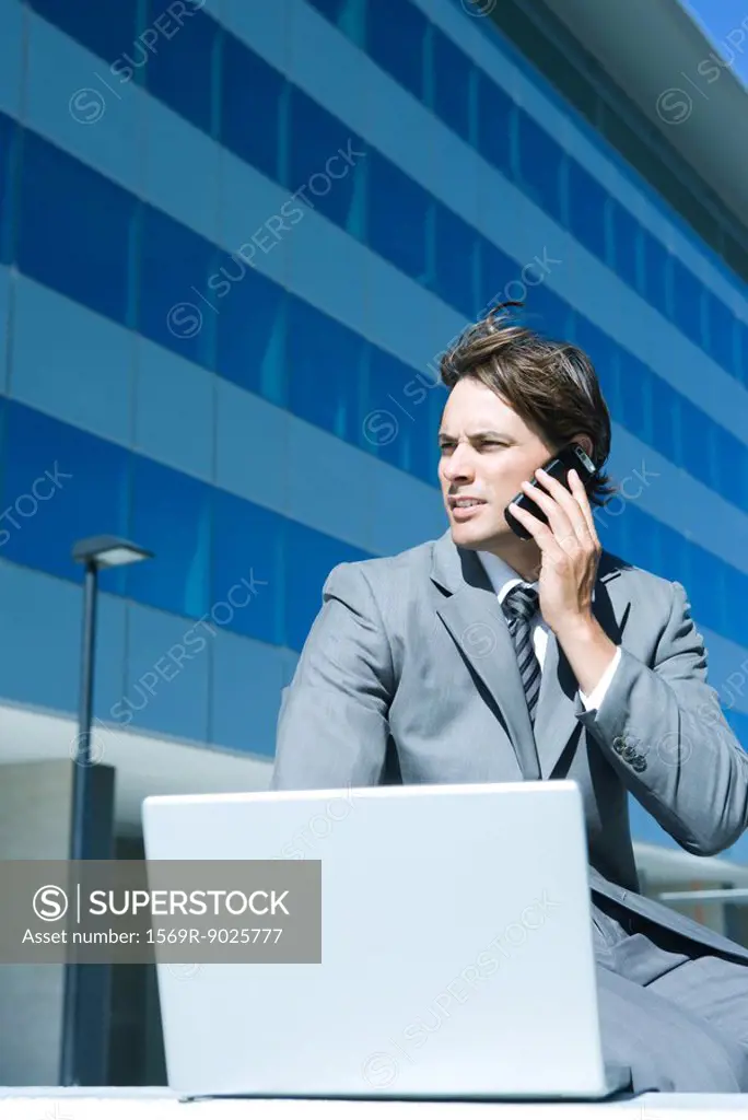 Young businessman sitting outdoors, using laptop and cell phone, looking away