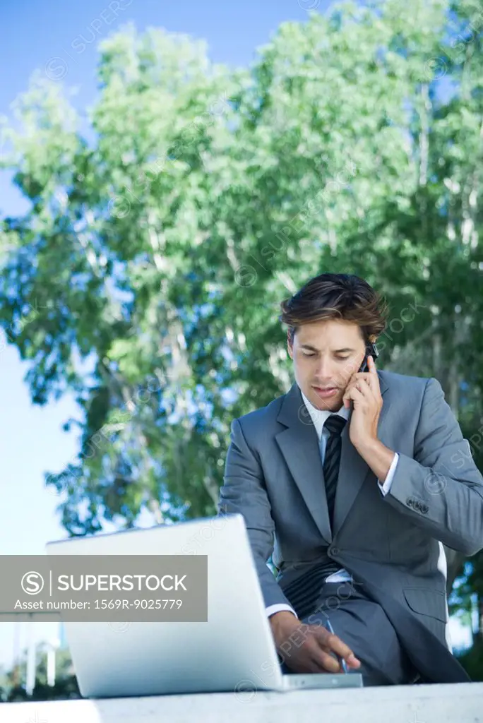 Young businessman sitting outdoors using cell phone and laptop computer, writing