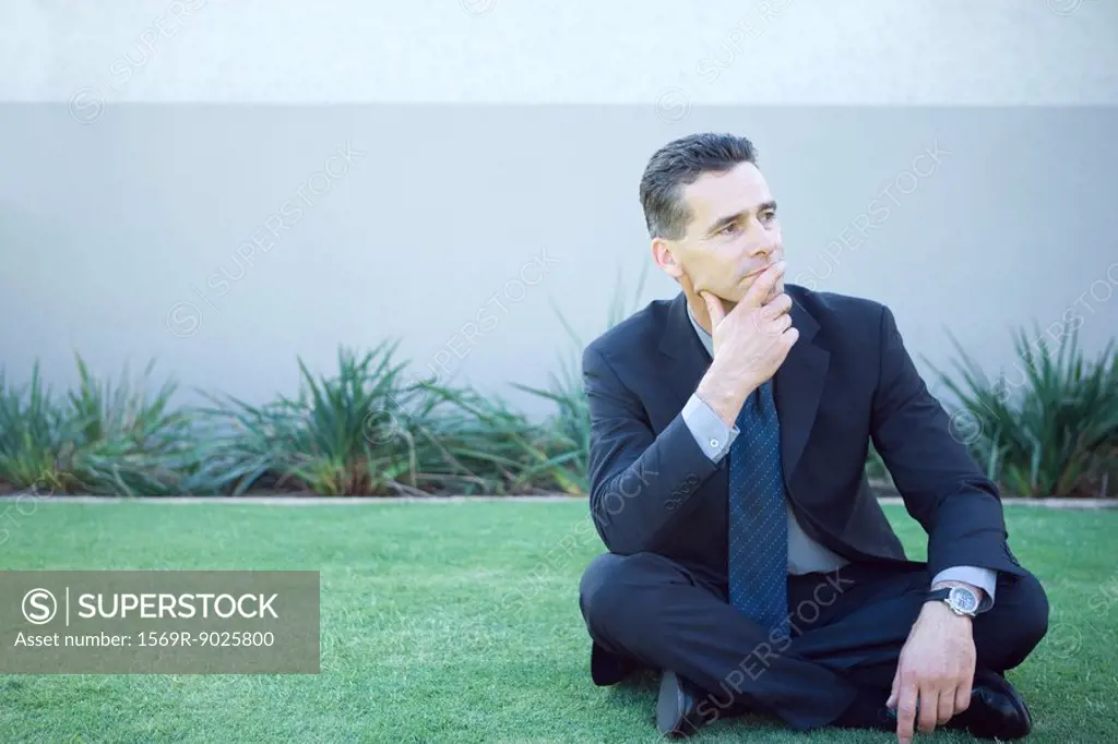 Businessman sitting on the ground outdoors, looking away, finger on lips