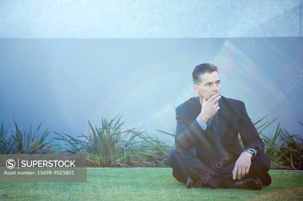Businessman sitting on the ground outdoors, looking away, finger on lips, lens flare