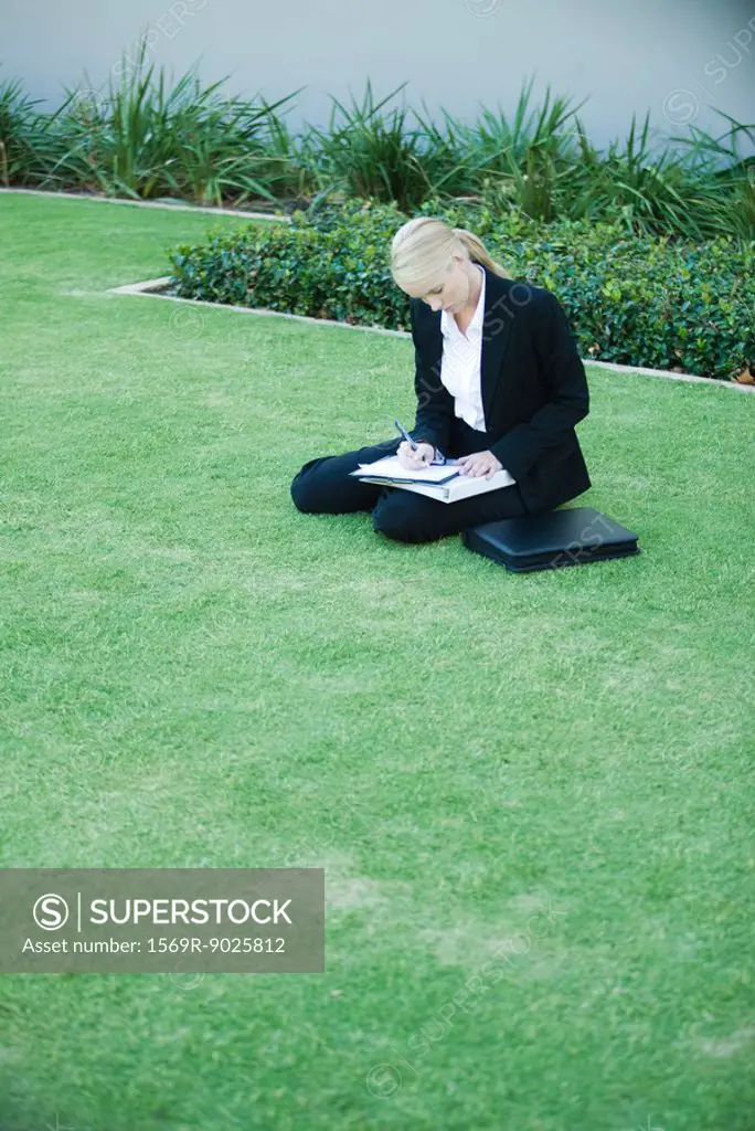 Young businesswoman sitting on the ground outdoors, writing on clipboard
