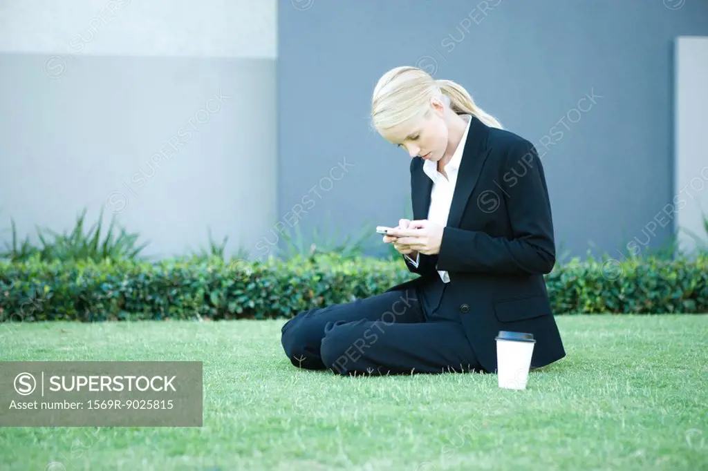 Young businesswoman sitting on the ground outdoors, looking at cell phone, hot beverage