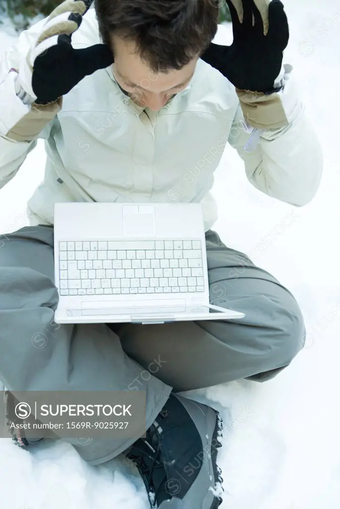 Young man sitting on the ground in snow, holding laptop computer on lap, hands raised