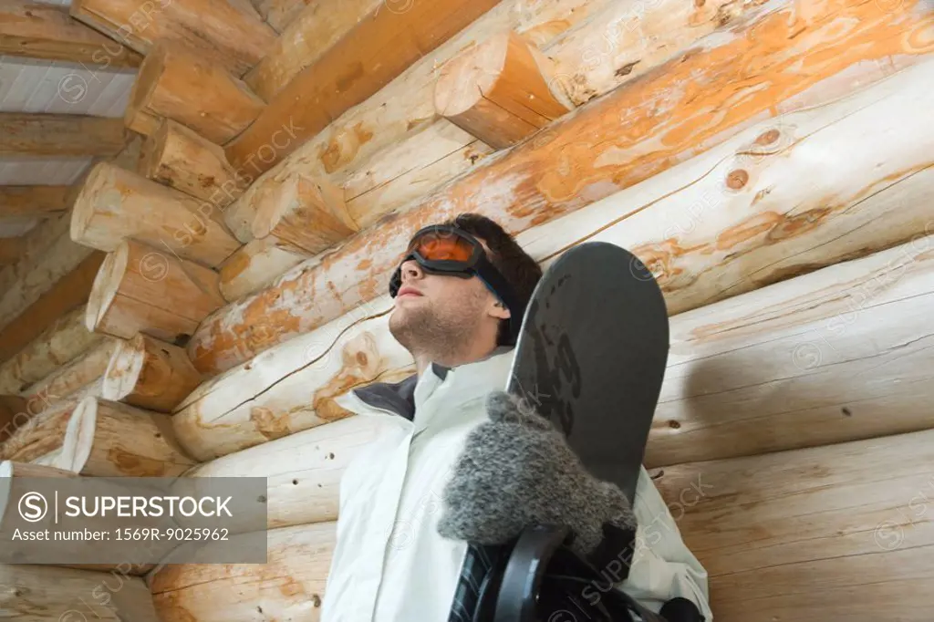 Young man with snowboard, leaning against log cabin, low angle view