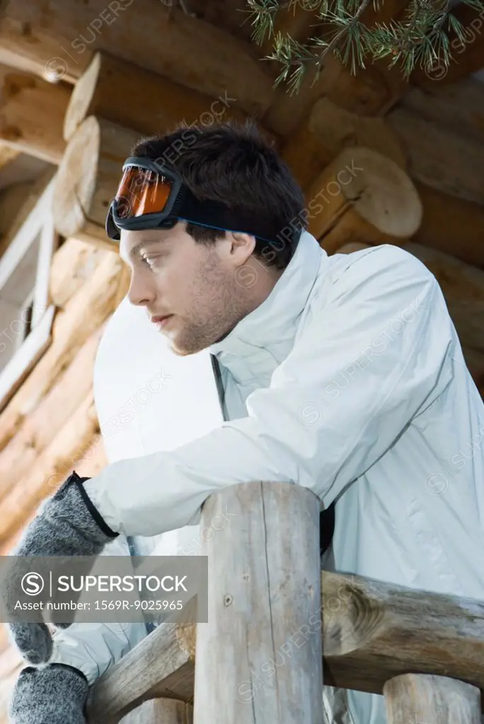 Young man in ski gear standing against deck of log cabin, leaning on rail