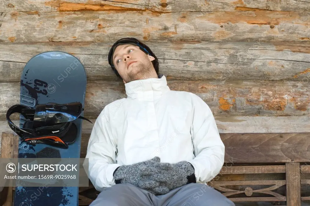 Young man sitting on bench next to snowboard, looking away