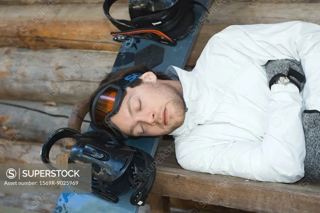 Young man lying on bench, resting head on snowboard, eyes closed