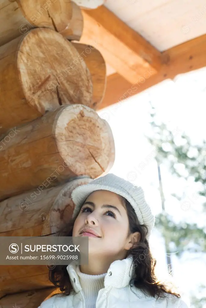 Teenage girl looking up, standing next to log cabin, portrait