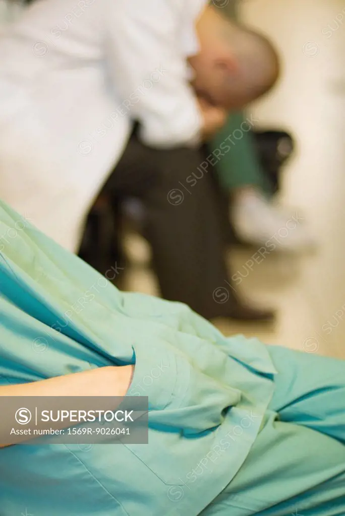 Medical worker with hands in pockets, doctor with head down in background, cropped view