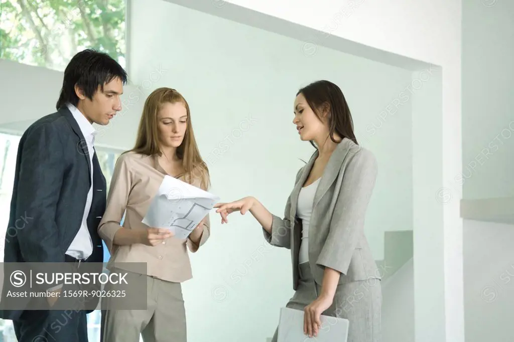 Female real estate agent standing in empty home interior with young couple, discussing blueprints