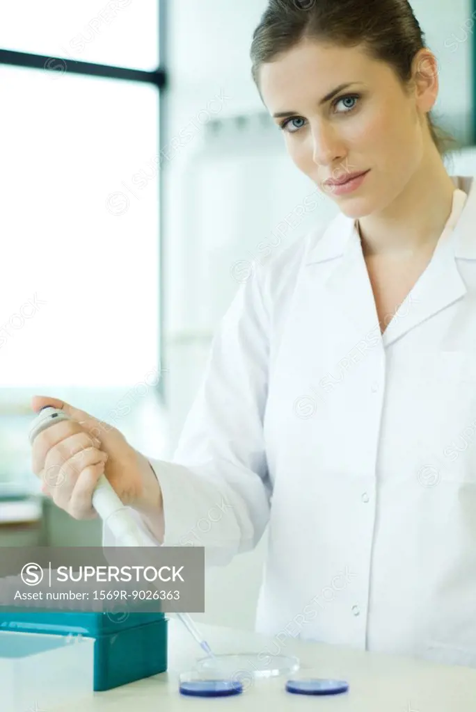 Young woman working in scientific laboratory, holding dropper over Petri dish, looking at camera