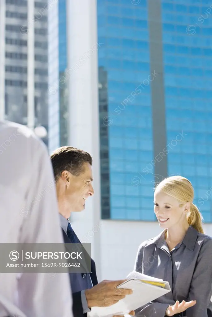 Business associates talking about file outdoors, smiling, office building in background