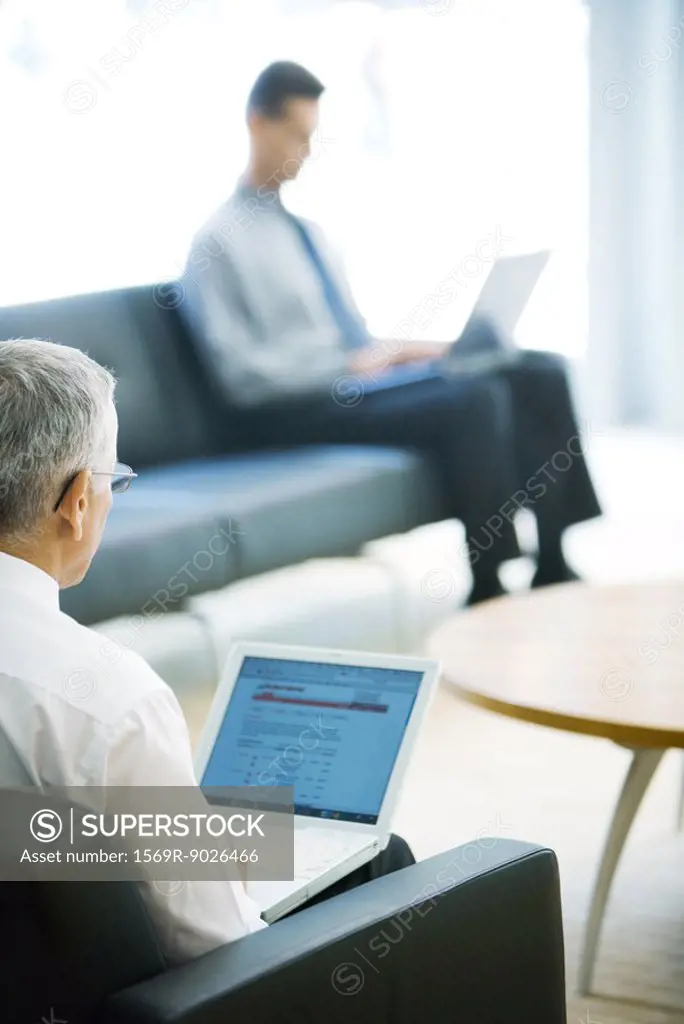 Two businessmen using laptops in lobby, focus on mature man in foreground