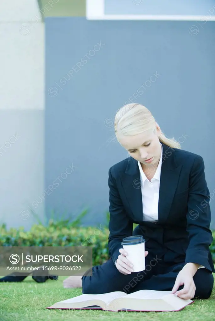 Businesswoman sitting on grass reading book, holding coffee cup