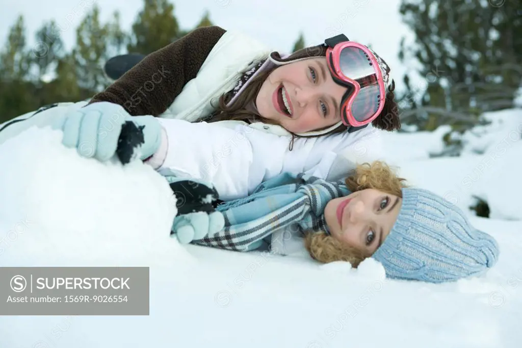 Two young friends reclining in snow, smiling at camera, one on top of the other