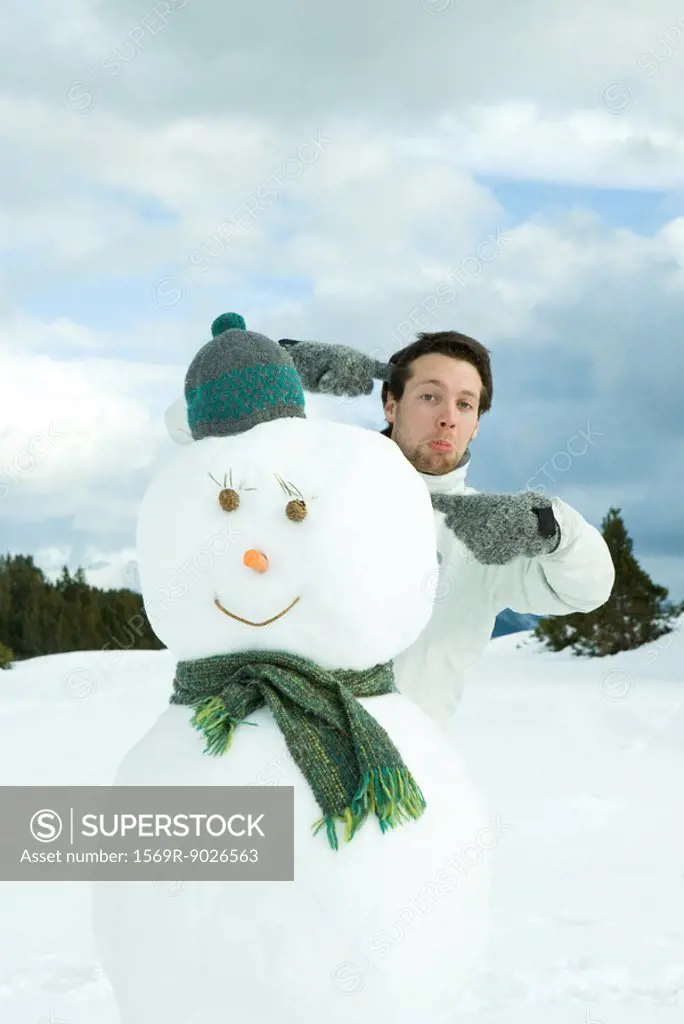 Young man standing behind snowman, pointing, making faces at camera