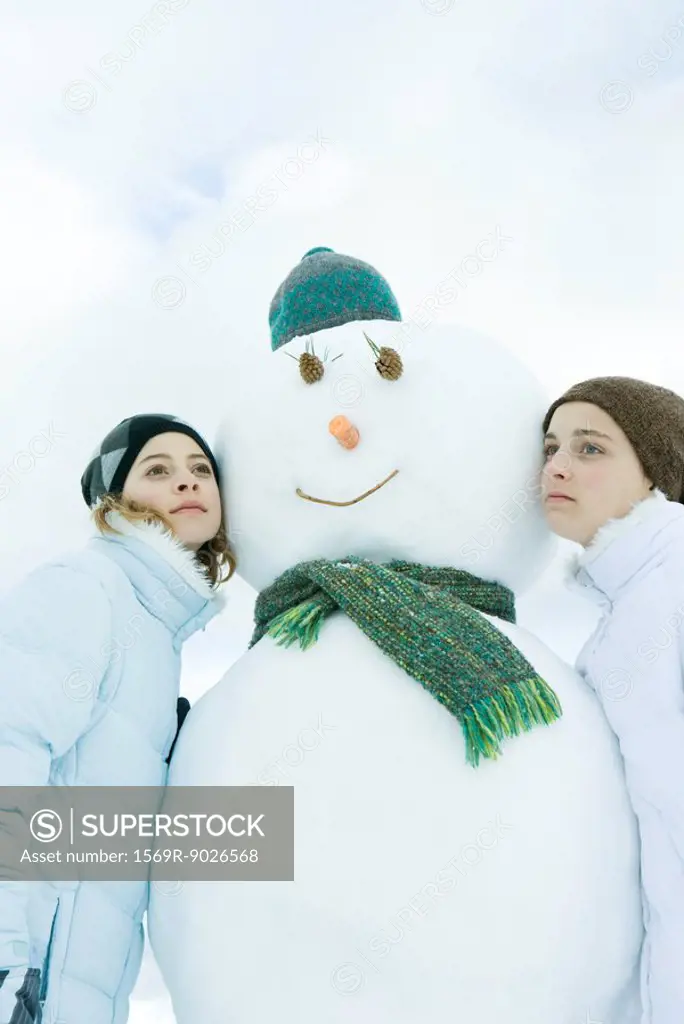 Two young friends leaning against snowman, looking away, portrait
