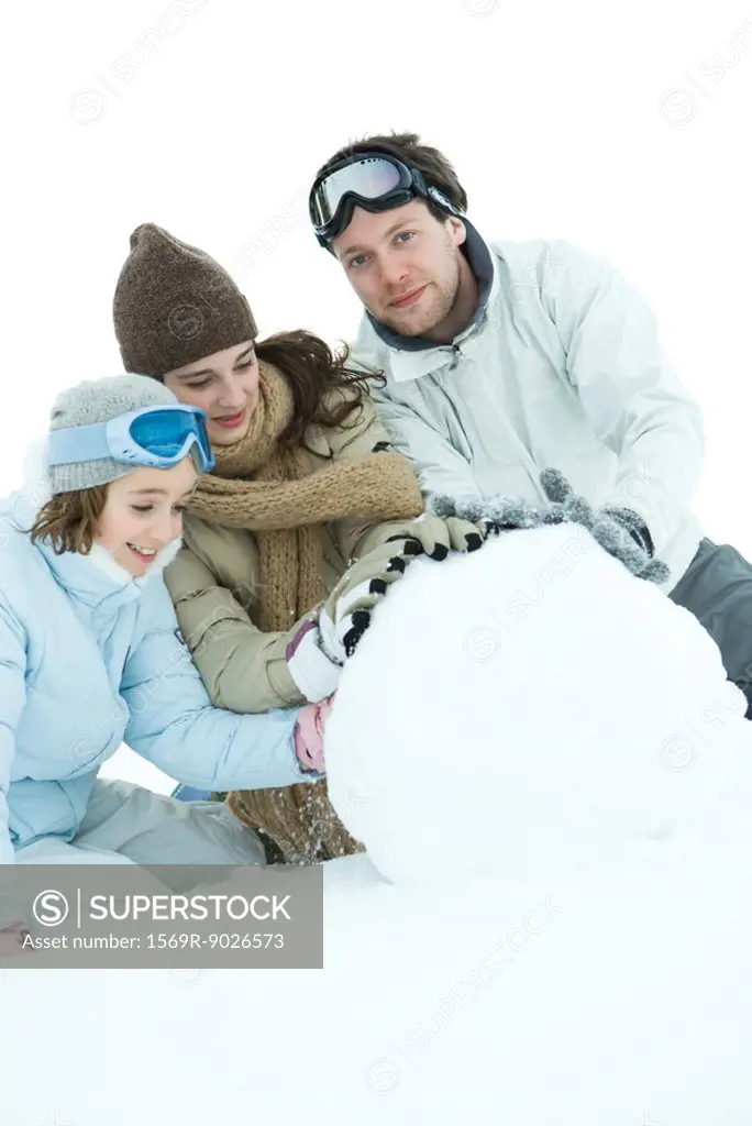 Three young friends making snowball, dressed in winter clothing, one looking at camera