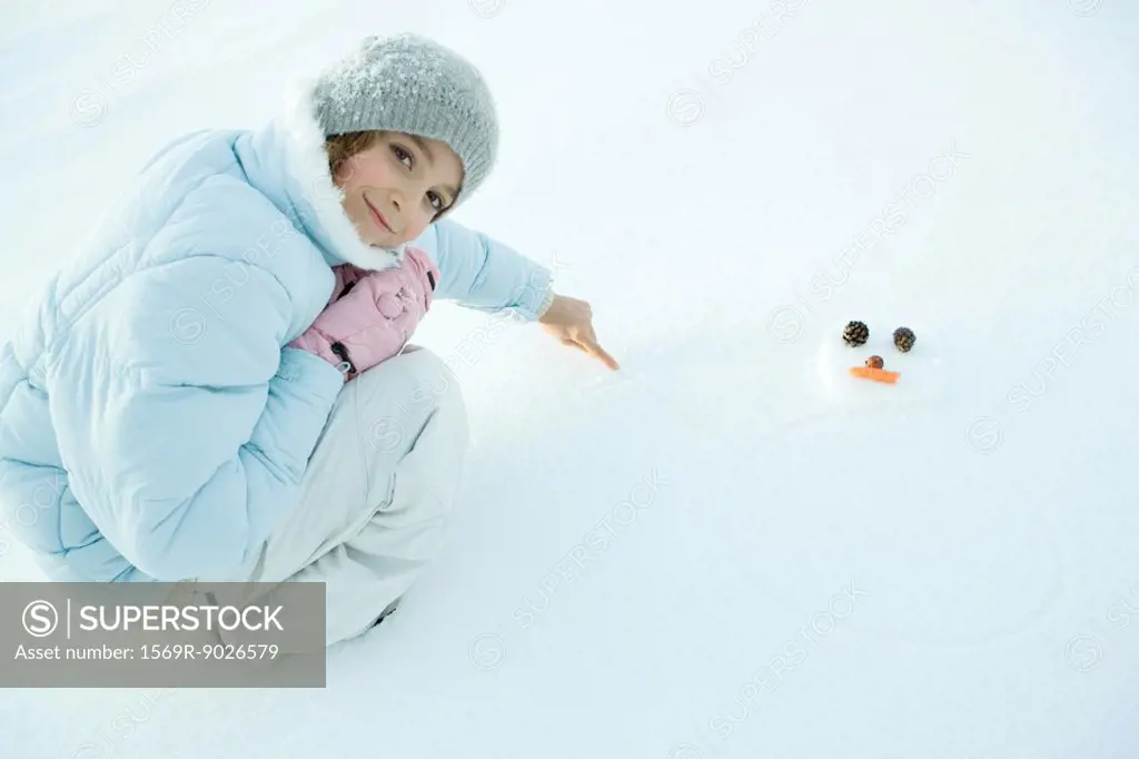 Preteen girl crouching in snow, drawing snowman, smiling at camera