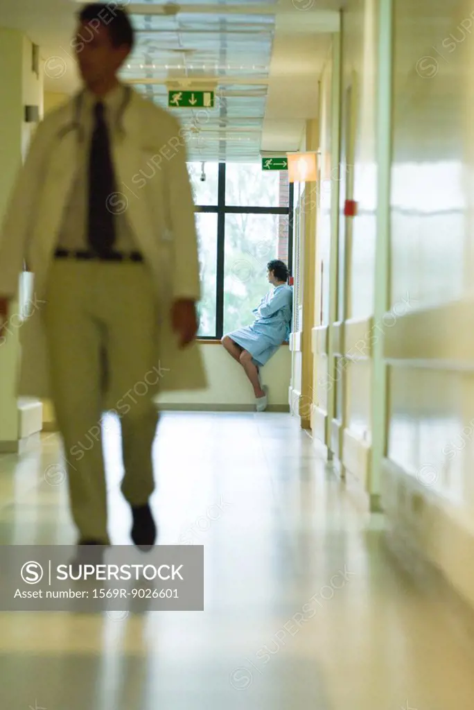 Man sitting, looking out window, wearing hospital gown, doctor walking in foreground