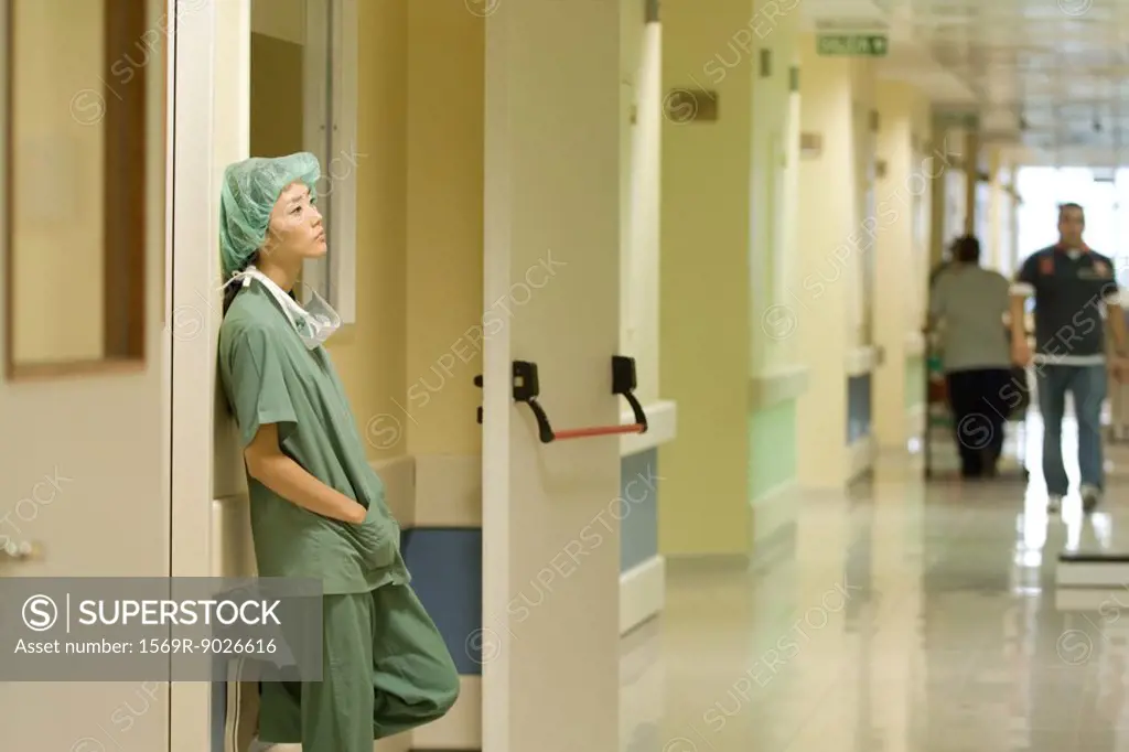 Female nurse in hospital, leaning against wall with hands in pockets, side view