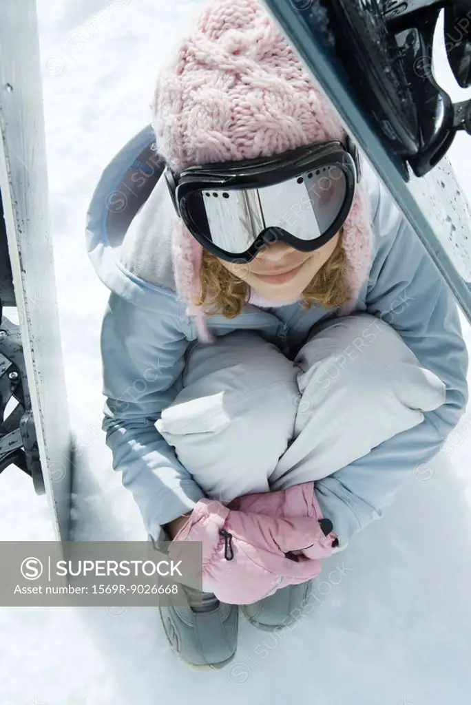 Preteen girl sitting on snow underneath propped up snowboards, high angle view