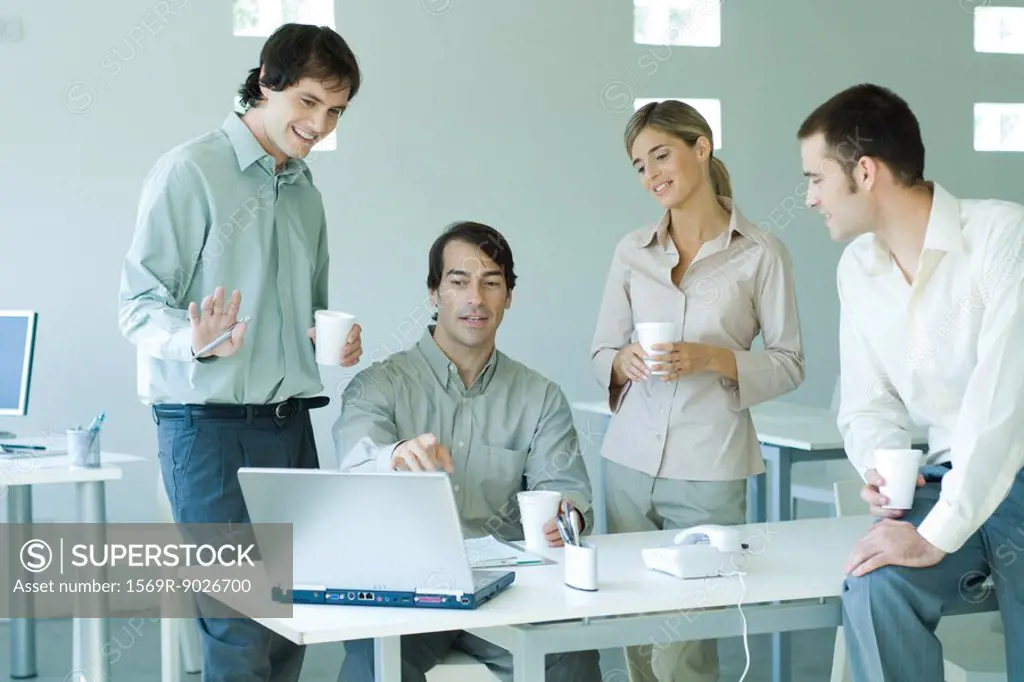 Four business associates in office, looking at laptop computer, holding coffee cups