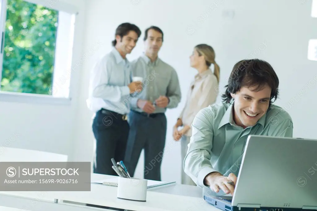 Businessman in office, using laptop computer, smiling, associates in background