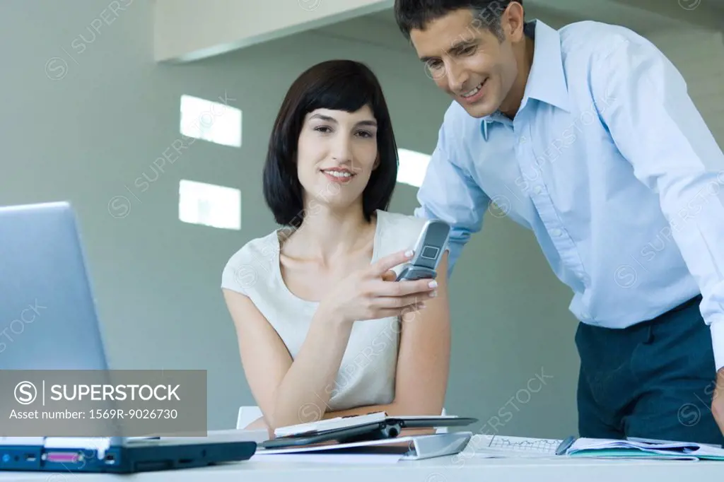 Young businesswoman holding cell phone, smiling at camera, businessman looking over shoulder