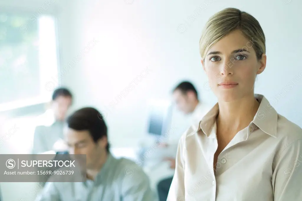 Young businesswoman looking at camera, head and shoulders, associates in background
