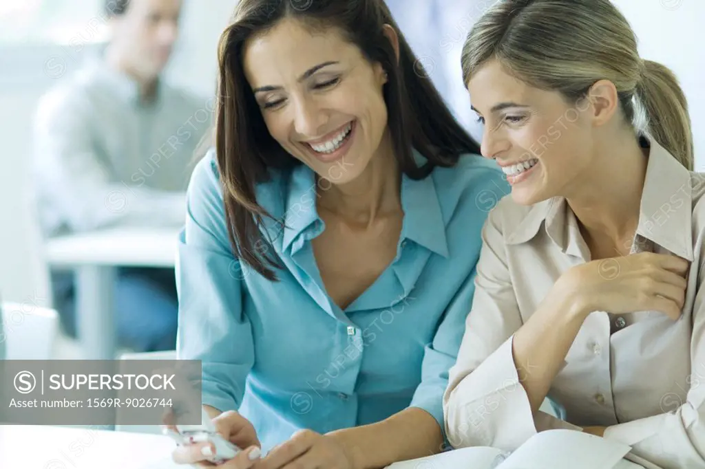 Two businesswomen looking at cell phone, smiling, waist up