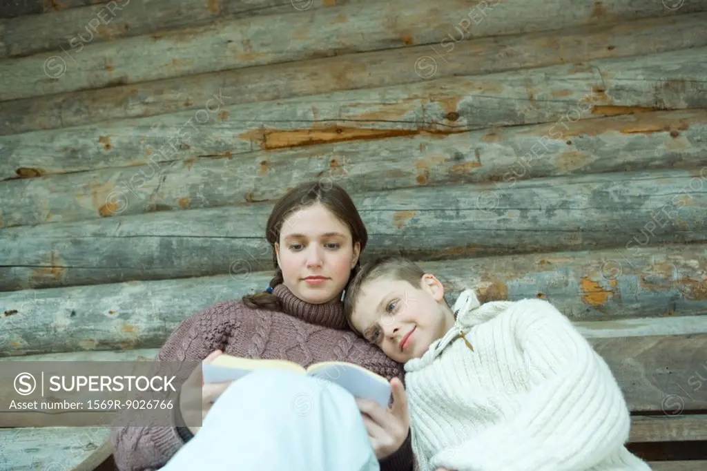Teenage girl reading book, little brother resting head on her shoulder