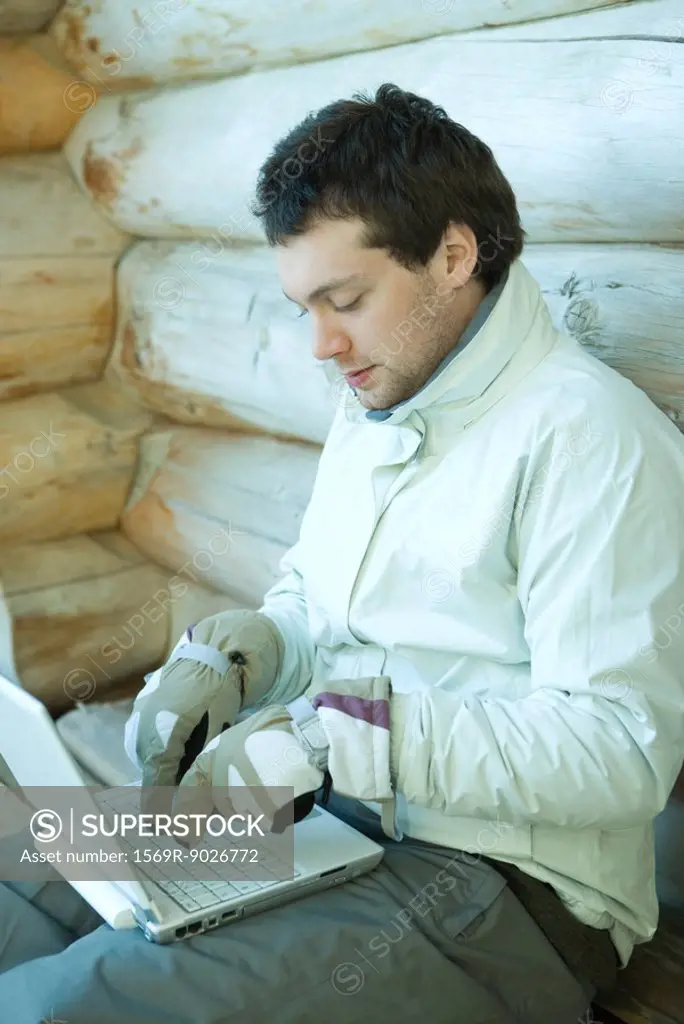 Young man using laptop computer, dressed in winter clothing, typing with gloves