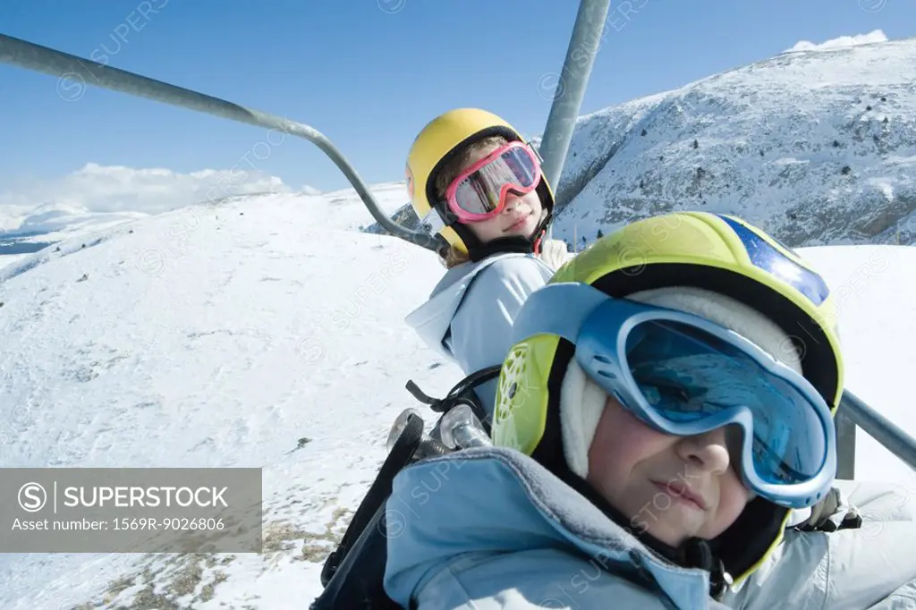 Two young friends on ski lift, both wearing ski goggles, one looking at camera