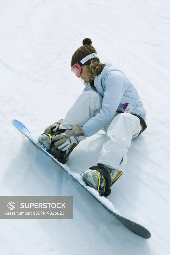 Teenage girl sitting on the ground, putting on snowboard, full length